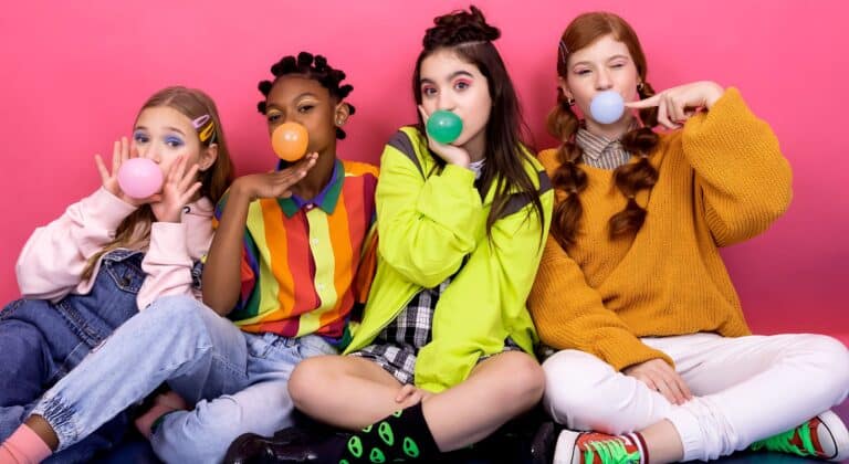 Four young girls blowing colorful bubblegum bubbles against a bright pink background, illustrating why colorful melatonin childrens gummies must be stored safely away from kids who confuse them with treats.